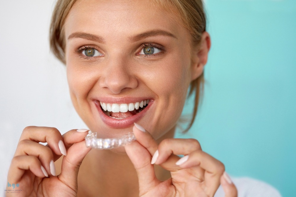 A blond girl smiling and holding Invisalign clear aligner close to her teeth, wondering about Invisalign cost in Charlottesville, VA
